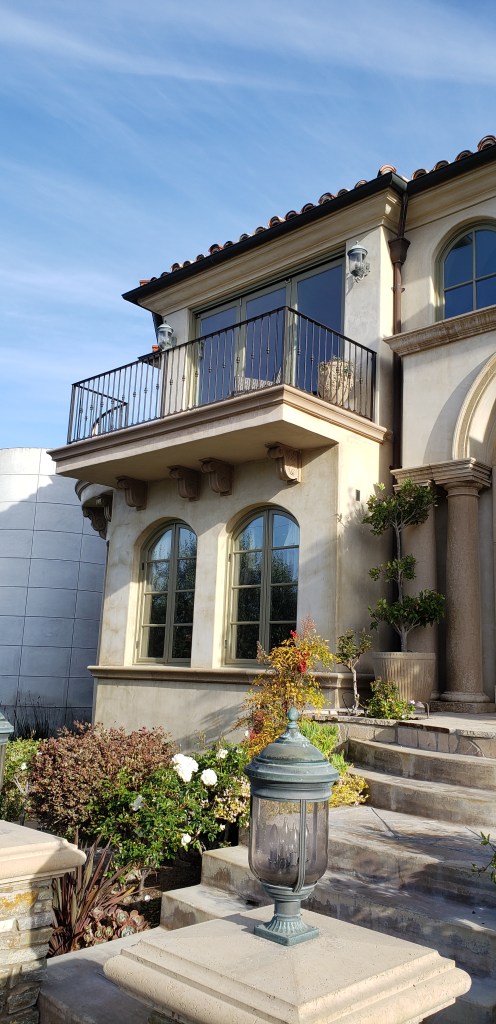 The image showcases a stunning two-story home with Mediterranean-inspired architecture. The upper level features a small balcony with a wrought iron railing, supported by ornate corbels that add a refined touch to the exterior. Below, arched windows contribute to the home’s classic and sophisticated aesthetic. The stone steps leading up to the house, paired with elegant lanterns and a carefully landscaped garden, create a warm and inviting atmosphere.